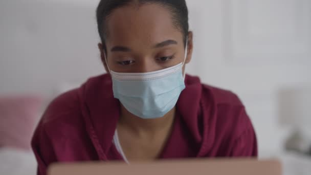 Nahaufnahme einer jungen konzentrierten Frau mit Covid-Gesichtsmaske, die auf ihrem Laptop im Bett im Internet surft. Porträt eines Afroamerikaners über die Pandemie. — Stockvideo