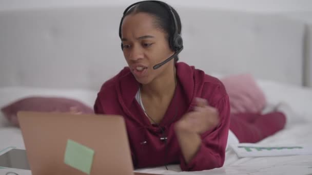 Retrato de la nerviosa joven afroamericana discutiendo en video chat acostada en la cama en la oficina del hogar. Gerente estresado insatisfecho peleando en conferencia virtual. Conflicto y trabajo remoto. — Vídeos de Stock