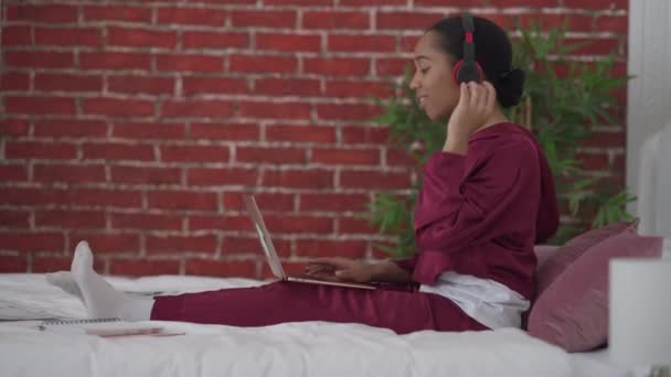 Side view vidám fiatal női szabadúszó élvezi a zenét a fejhallgató üzenetküldés online laptop a hálószobában. Széles lövés mosolygós gyönyörű afro-amerikai nő gépelés billentyűzeten tánc. — Stock videók