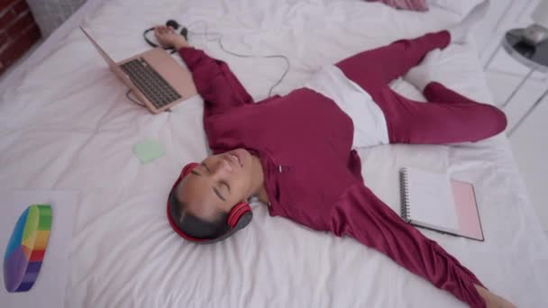 Joven mujer afroamericana satisfecha en pijamas y auriculares que vuelven a la cama riendo. Retrato de vista superior de alegre feliz hermosa freelancer disfrutando de trabajo remoto en casa oficina. — Vídeos de Stock