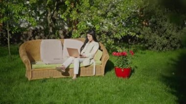 Mutlu, kendine güvenen Asyalı bir kadının güneşli bahçede oturmuş dizüstü bilgisayarında daktilo yazarken son derece geniş bir görüntüsü. Baharın arka bahçesinde internette mesajlaşan neşeli, güzel iş kadınının portresi..