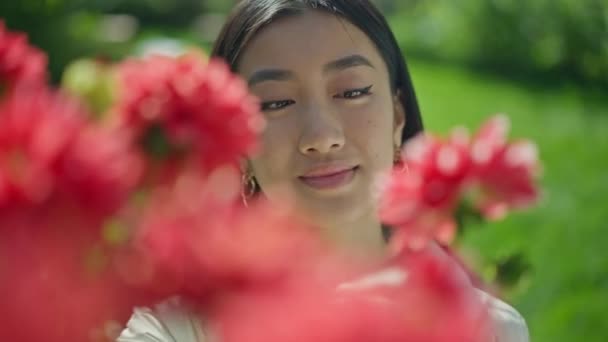 Cara de cerca de la joven mujer asiática feliz con ojos marrones detrás de flores rojas en flor en el jardín soleado. Retrato de la cabeza de florista hermosa delgada satisfecha jardinero admirando flor al aire libre. — Vídeos de Stock