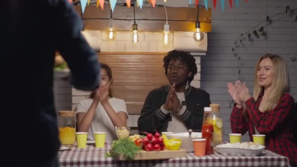 Grupo multiétnico de personas aplaudiendo como hombre sonriente trayendo sabrosas hamburguesas a la mesa de picnic. Felices amigos relajados y familiares disfrutando de la comida al aire libre en el jardín de verano por la noche. — Vídeo de stock