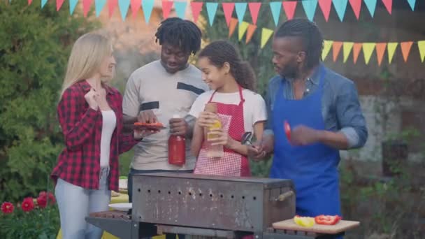 Middle shot of cheerful laughing group of multinational young people chatting on picnic outdoors. Happy smiling Caucasian woman African American men and teen girl talking cooking bbq in summer garden. — Stock Video