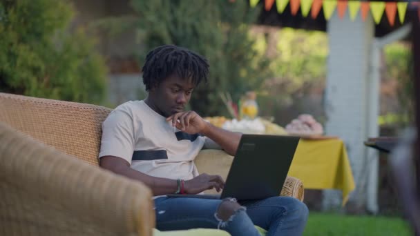 Absorbed young African American man typing on laptop keyboard sitting on couch in summer garden. Portrait of confident professional expert working online on backyard messaging. Remote working concept. — Stock Video