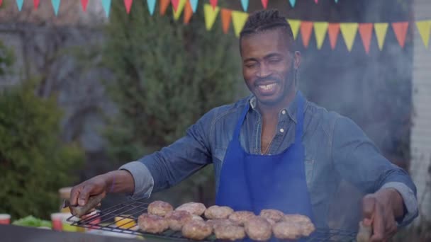 Retrato de feliz sorrindo homem afro-americano posando com hambúrgueres na grade de churrasco ao ar livre. Joyful jovem chef bonito com sorriso de dente se gabando carne grelhada assada no churrasco piquenique. — Vídeo de Stock