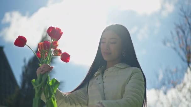 Jovem sorrindo mulher asiática cortando hastes de flores com tesoura em pé à luz do sol com céu nublado azul primavera verão no fundo, Retrato de florista feminino sorridente fazendo buquê de tulipas no jardim. — Vídeo de Stock
