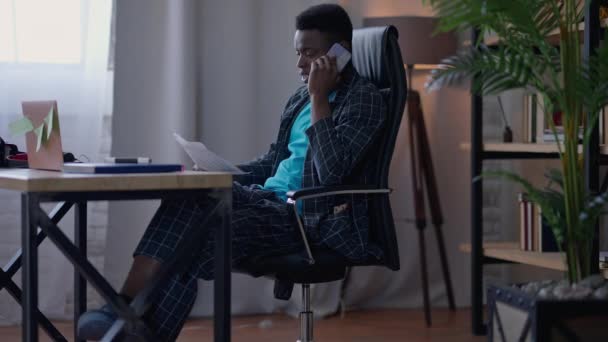 Amplio tiro de confianza joven consultor de negocios guapo hablando en el teléfono inteligente analizar gráfico de negocios en la mano. Vista lateral del hombre afroamericano en pijama socio convincente de la oficina en casa. — Vídeo de stock