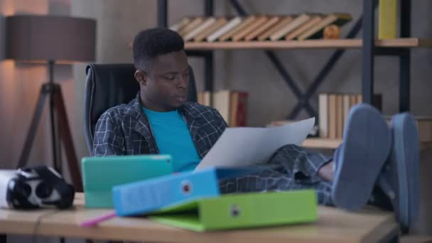 自宅の事務所で書類を調べるテーブルの上に足でパジャマを着たアフリカ系アメリカ人の若者の大規模なショット。屋内のリビングルームでスタートアッププロジェクトを分析するハンサムな自信のあるビジネスマン. — ストック動画