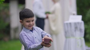 Yüzük kutusu gösteren heyecanlı Orta Doğulu çocuk gülümseyen kameraya bakıyor. Evlilik töreninde dışarıda takılarla poz veren neşeli çocuk portresi. Bulanık çift evleniyor.