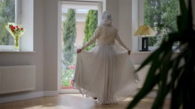 Gelinlik giymiş, tesettürlü ve tesettürlü genç Orta Doğulu bir kadının sabahları mihraba doğru yürüdüğünü gösteren geniş bir bakış açısı. Kendine güveni tam mutlu gelin evde hazırlanıyor..