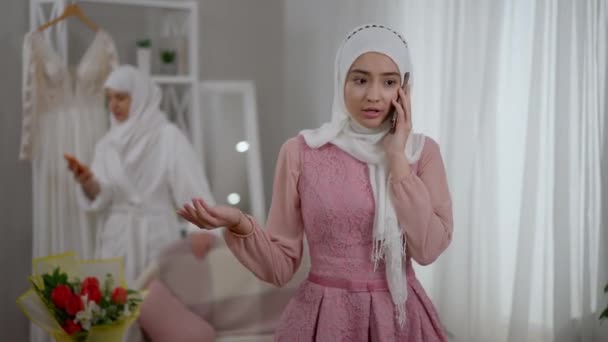 Ansiosa joven dama de honor de Oriente Medio hablando por teléfono con la novia preocupada en el vestido de novia en el fondo. Retrato de mujer musulmana moderna y segura en hiyab discutiendo los preparativos de la boda. — Vídeos de Stock