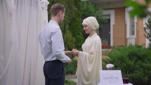 Piękna, bliskowschodnia panna młoda wkładająca pierścionek na palec białego mężczyzny uśmiechającego się. Portret kochającej kobiety w sukni ślubnej i hidżabie biorącej ślub na świeżym powietrzu. — Wideo stockowe