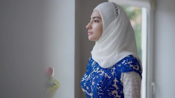 Noiva séria e confiante do Oriente Médio em hijab e vestido de noiva olhando para fora da janela se transformando em câmera. Retrato de jovem bonita magro com olhos castanhos pensando dentro de casa. — Vídeo de Stock