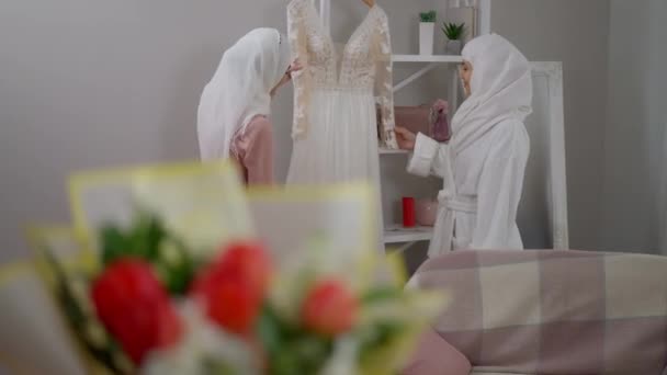 Brede opname van twee vrolijke lachende vrouwen in hijabs die de trouwjurk bewonderen en glimlachend praten en weglopen. Midden-Oosterse bruid en bruidsmeisje maken zich klaar in de ochtend binnen. — Stockvideo