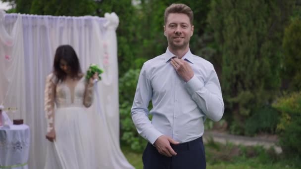 Magabiztos, büszke kaukázusi férfi pózol a házassági ceremónián, visszanéz a boldog közel-keleti nőre, aki a ruhát bámulja a háttérben. Portré mosolygós jóképű vőlegény a kertben szabadban. — Stock videók