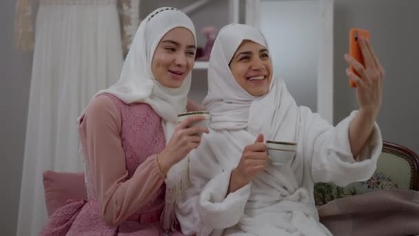 Lachen vrolijke moslimbruid en bruidsmeisje praten op smartphone streaming in sociale media op huwelijksochtend. Vrolijke opgewonden Midden-Oosten vrouwen in hijabs opscheppen online met theekoppen. — Stockvideo