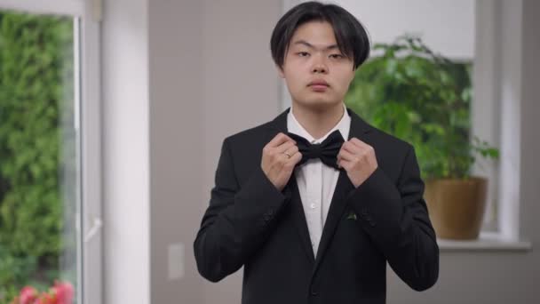 Mirror POV of Asian handsome groom trying on bow tie smiling. Portrait of happy man in elegant wedding suit standing indoors getting dressed. Marriage and confidence concept. — Stock Video