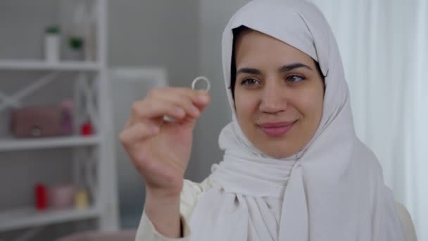 Linda jovem mulher do Oriente Médio com olhos de avelã em hijab branco admirando anel de noivado na mão. Retrato de noiva bonita confiante feliz no dia do casamento dentro de casa. Casamento e felicidade. — Vídeo de Stock