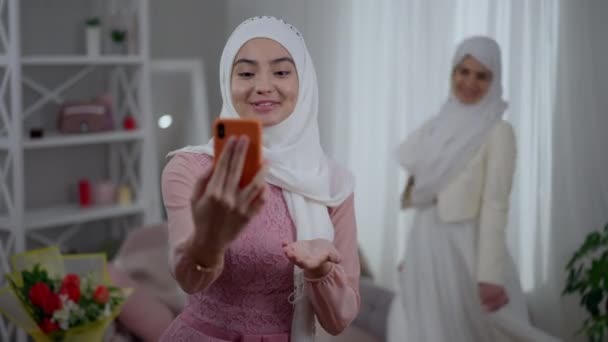 Moderno Médio Oriente dama de honra streaming on-line usando smartphone falando e sorrindo como noiva feliz girando em vestido de noiva no fundo. Mulher encantadora apoiando amigo se casar. — Vídeo de Stock