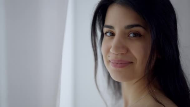 Hovedbillede portræt af slank ung smuk mellemøstlig kvinde kigger ud af vinduet og ser på kameraet. Close-up ansigt af smuk selvsikker dame poserer indendørs om morgenen. – Stock-video
