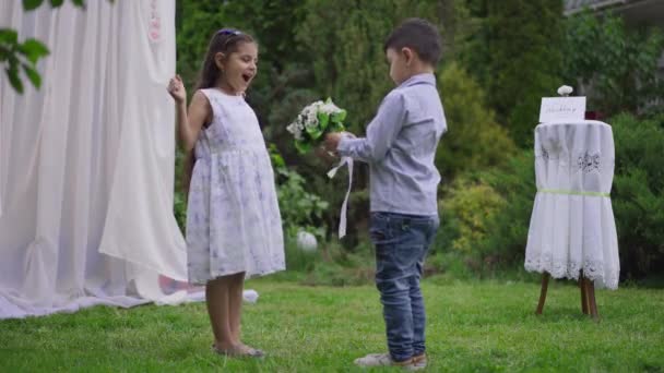 Garota bonita Oriente Médio animado recebendo buquê de casamento de menino bonito no altar. Retrato de tiro largo de crianças positivas desfrutando de cerimônia de casamento no jardim da primavera ao ar livre rindo se divertindo. — Vídeo de Stock