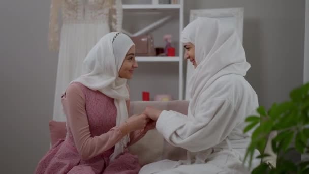Вид сбоку портрет улыбающейся ближневосточной подружки невесты, держащей за руки красивую стройную невесту, разговаривающую и обнимающую подругу. Две молодые счастливые мусульманки в помещении в день свадьбы. Поддержка и поддержка. — стоковое видео