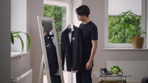 Jonge bruidegom probeert een trouwpak aan en kijkt in de spiegel in de woonkamer. Zijaanzicht portret van Aziatische man bewonderende reflectie holding hanger met elegante kostuum met boutonniere. Voorbereiding op het huwelijk. — Stockvideo