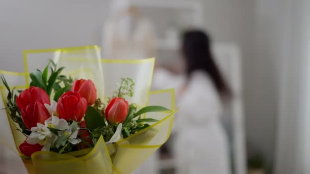 Primer plano del ramo de flores en el interior con novia joven borrosa que toma el vestido de novia y se va al fondo. Mujer irreconocible preparándose por la mañana en el día de la boda. — Vídeo de stock