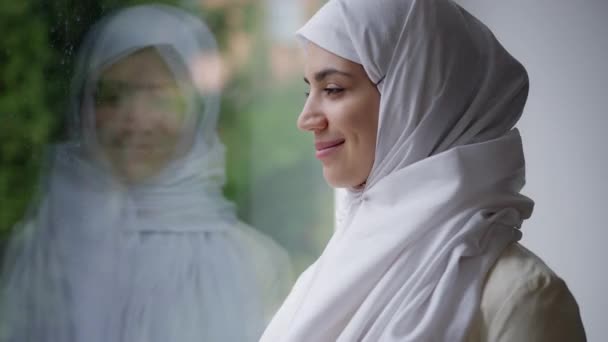 Junge schöne Frau aus dem Nahen Osten mit charmantem Lächeln, die lächelnd in die Kamera blickt. Porträt einer glücklichen selbstbewussten Nahostbraut im weißen Hijab, die am Fenster posiert. — Stockvideo