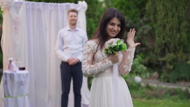 Портрет веселої нареченої з Близького Сходу, яка хвалиться обручкою на пальцях, дивиться на фотоапарат, танцюючи, як розмитий усміхається кавказький наречений стоїть на задньому плані. Любов і збудження. — стокове відео