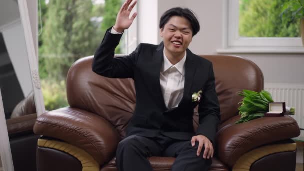 Fröhlicher frischgebackener asiatischer Mann mit zahmem Lächeln, der in die Kamera winkt und Ring an Finger zeigt. Porträt eines glücklichen, aufgeregten Bräutigams, der lächelnd auf einem Sessel sitzt. Video-Chat. — Stockvideo