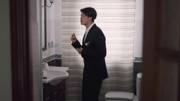 Widok z boku portret szczęśliwy podekscytowany azjatycki pan młody w garniturze ślubnym taniec w łazience patrząc na lustro śpiew. Radosny młodzieniec radujący się w dniu ślubu przygotowuje się. Małżeństwo i szczęście. — Wideo stockowe