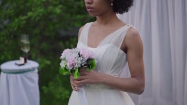 Beyaz elbiseli Afro-Amerikalı genç bir kadın mihrapta düğün buketi tutuyor. Zarif, ince, güzel gelin bahar bahçesinde evleniyor..