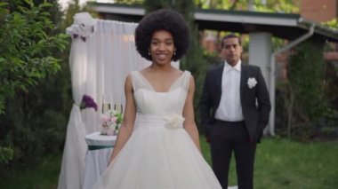 Mutlu Afro-Amerikan çekici gelinin portresi. Gelinlik içinde, açık havada, yaz bahçesinde poz veriyor. Beyazlar içindeki muhteşem kadın zarif damada bakıp kameraya gülümsüyor..