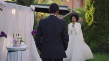 Gelinlik giymiş kendine güvenen damadın mihrapta dikilirken beyaz elbiseli bulanık gülümseyen gelinin partnerine ağır çekimde yürümesi. Mutlu Afrikalı Amerikalı çift açık havada evleniyor..
