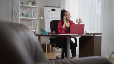 Afro-Amerikan genç bir kadının sabah kapuçinosu içerken portresi dahili ofisteki masada oturmuş akıllı telefondan konuşuyor. Gülümseyen güzel milenyum planlama zamanı kapalı alanda çalışma zamanı.