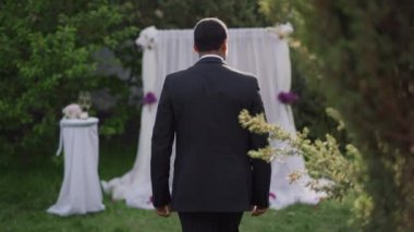 Genç, zarif Afrikalı Amerikalı damat ağır çekimde düğün mihrabına yürüyor misafirlere dönüyor ve el sallıyor. Siyah kostümlü mutlu bir adamın dışarıda evlenmesinin portresi..