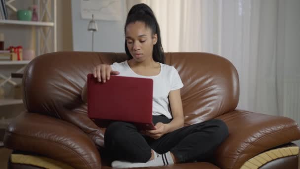 Retrato de una joven mujer confiada cerrando la computadora portátil de pie desde un cómodo sillón y saliendo de la oficina en casa. Slim hermosa dama afroamericana al final del tiempo de trabajo alejándose en cámara lenta — Vídeos de Stock