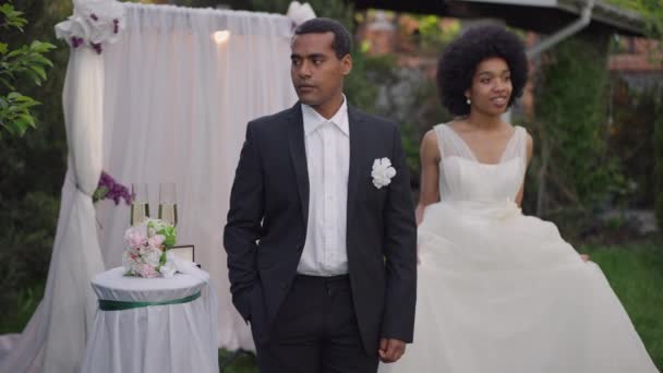 Guapo novio confiado en traje de pie al aire libre como novia acercándose tomando la mano de la pareja mirando con amorosa expresión facial. Alegre sonriente afroamericano recién casados posando en ceremonia. — Vídeos de Stock