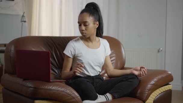 Joven encantadora mujer confiada girando en la lista de reproducción en el ordenador portátil sentado en la pose de loto en el sillón. Amplio retrato de yogui afroamericano femenino en la oficina del hogar en el trabajo remoto. Tranquilidad. — Vídeo de stock