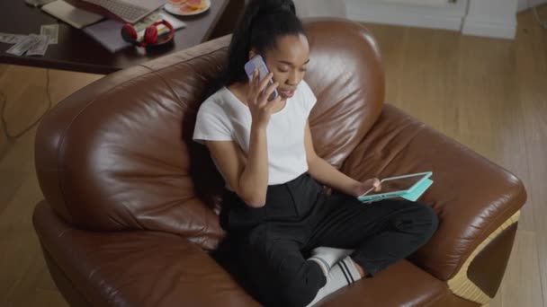 Hög vinkel syn på säker expert sitter på mysiga fåtölj i hemmakontoret talar i telefon. Brett skott av smal vacker afroamerikansk kvinna övertygande affärspartner i idé lönsamhet. — Stockvideo