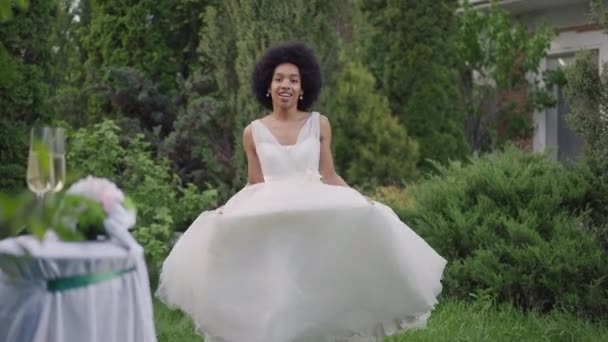 Amplio tiro de alegre novia afro-americana emocionada en vestido elegante blanco corriendo en cámara lenta en el prado verde del verano y girando. Retrato de mujer alegre casándose al aire libre. — Vídeos de Stock