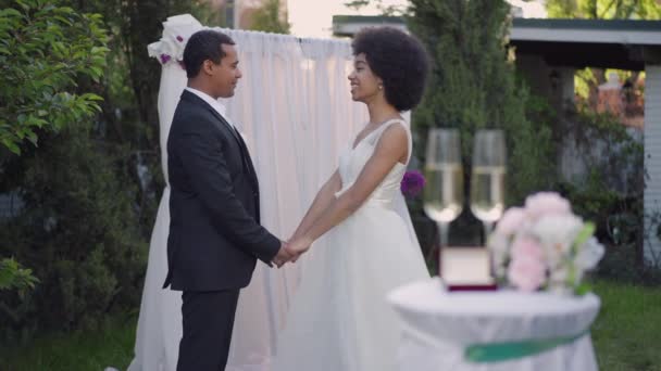 Zijaanzicht van vreugdevol koppel dat hand in hand aan altaar staat te praten in slow motion bij zonsondergang. Gelukkige glimlachende Afro-Amerikaanse bruid en bruidegom chatten buiten. Geluk en huwelijk. — Stockvideo