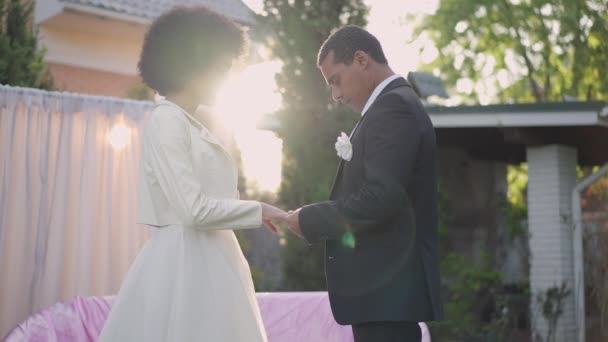 Pareja afroamericana joven y cariñosa parada bajo la luz del sol al aire libre tomados de la mano mirándose el uno al otro con amor. Retrato vista lateral de novio y novia cariñosos en rayos de sol en el jardín. — Vídeos de Stock