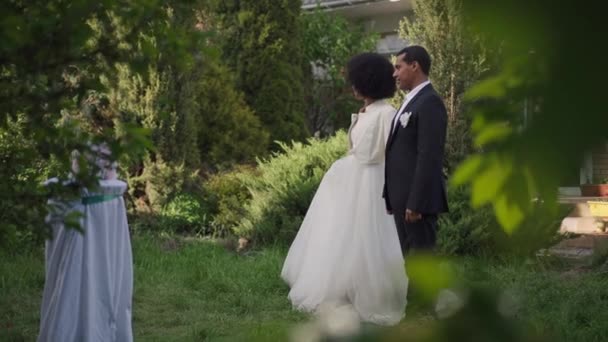 Vista lateral amplia toma de feliz pareja afroamericana de recién casados caminando en cámara lenta en el jardín de verano. Mujer hermosa sonriente y hombre guapo confiado paseando al aire libre en la ceremonia de matrimonio. — Vídeos de Stock