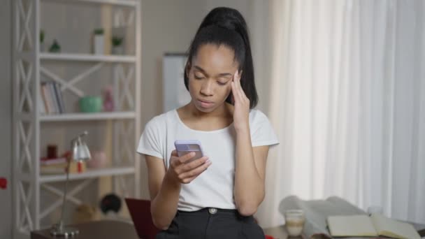 Troubled giovane donna con sintomi di emicrania messaggistica su smartphone sfregamento tempie. Ritratto di triste donna d'affari afroamericana malata che lavora online dall'ufficio di casa. Problemi sanitari. — Video Stock