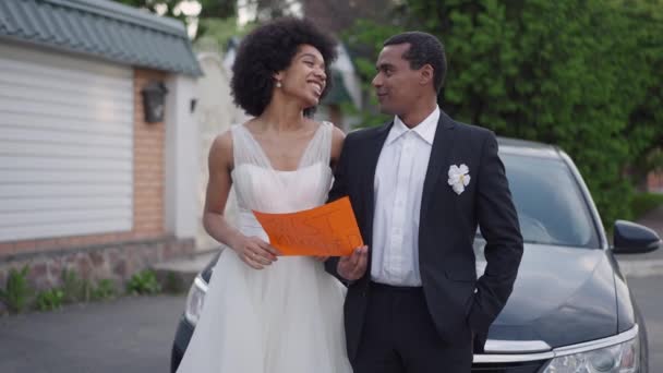 Feliz pareja joven afroamericana de pie en el coche sosteniendo el cartel de casados hablando sonriente. Los recién casados alegres en el vehículo salen para la luna de miel después de la ceremonia de boda. Romance y amor. — Vídeos de Stock