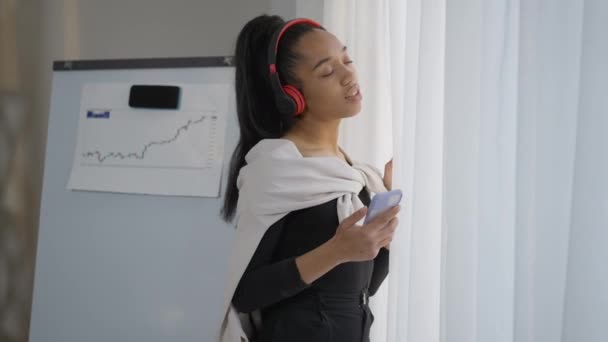 Retrato de vista lateral de la alegre mujer joven relajada en auriculares bailando en cámara lenta escuchando la lista de reproducción en el teléfono inteligente. Irresponsable descuidado empleado afroamericano en el Ministerio del Interior. — Vídeos de Stock