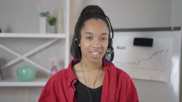 Vooraanzicht portret van vrolijke glimlachende Afro-Amerikaanse jonge vrouw in oortelefoons praten in virtuele conferentie. Web camera POV van slimme mooie business consultant conferencing vanuit kantoor. — Stockvideo
